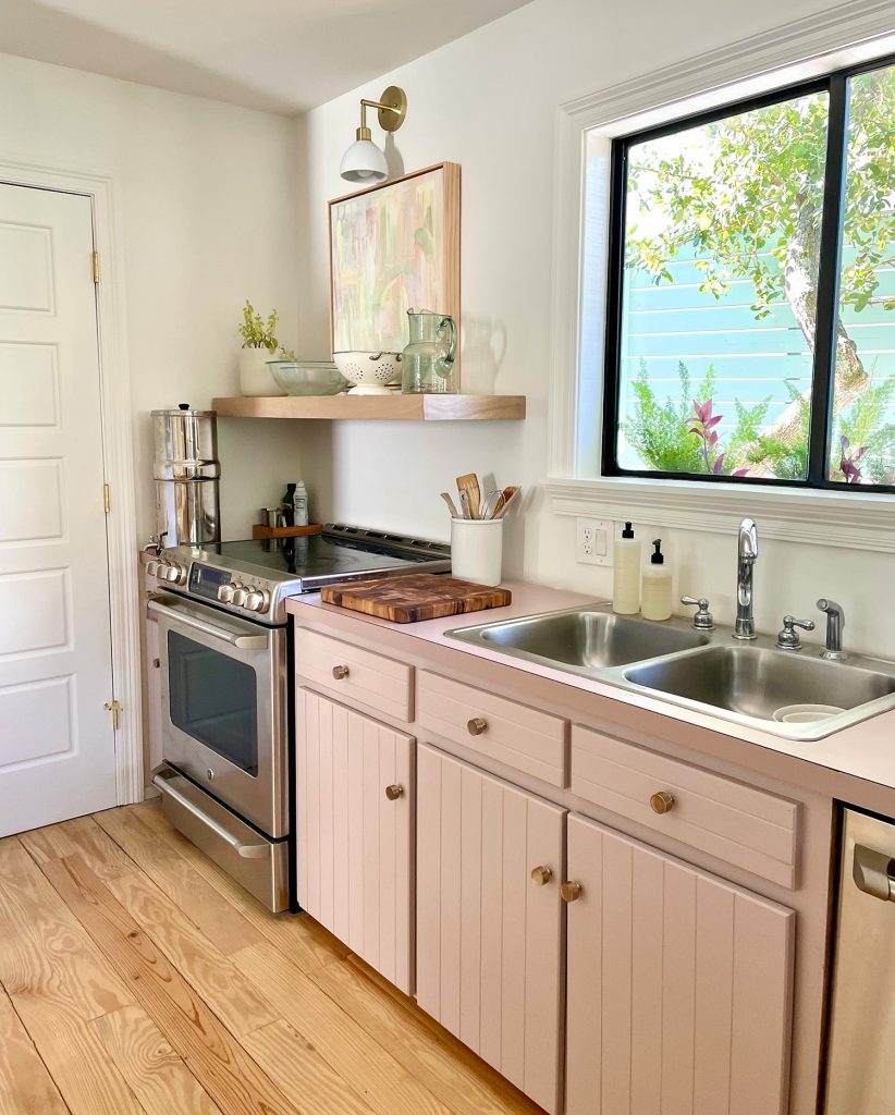 Beachy Kitchen With Mauve Pink Cabinets And Wood Floating Shelves With Mid Century Modern Lights