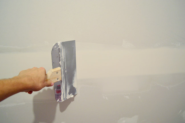 John dragging large drywall knife over seam in drywall to smooth mud