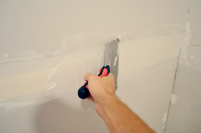 larger drywall knife being dragged across tape to apply another layer of mud